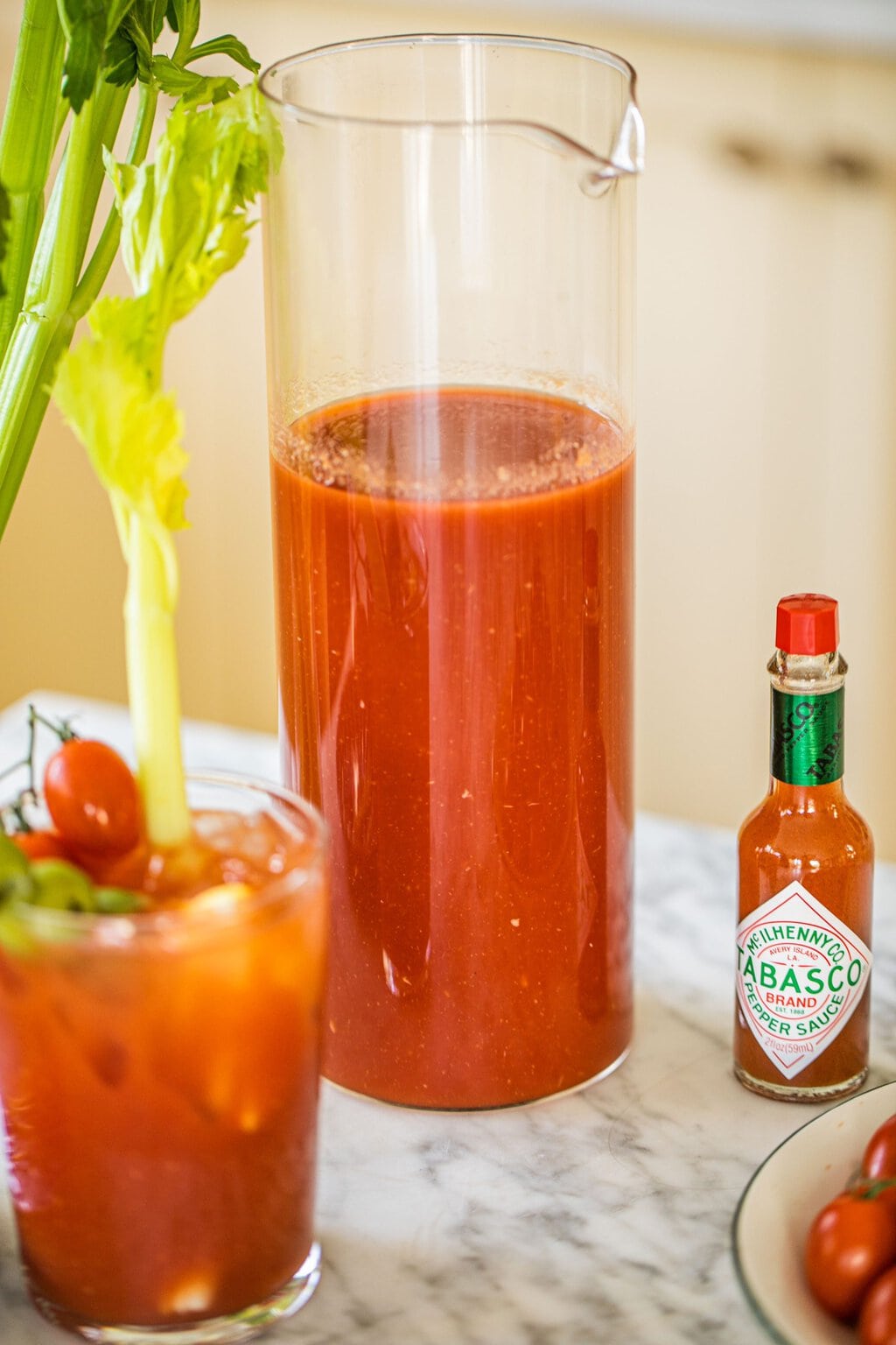 a pitcher of bloody mary mix beside tabasco sauce and with a glass of the cocktail and garnish in front