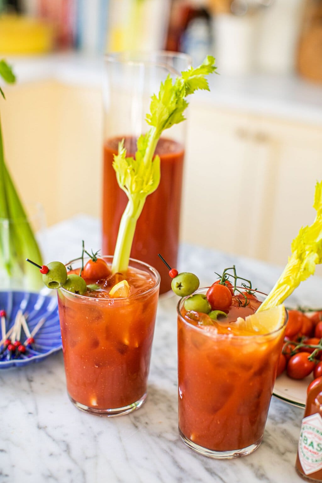 two glasses of bloody marys with celery, olives, and tomatoes as garnish and a large pitcher in the background