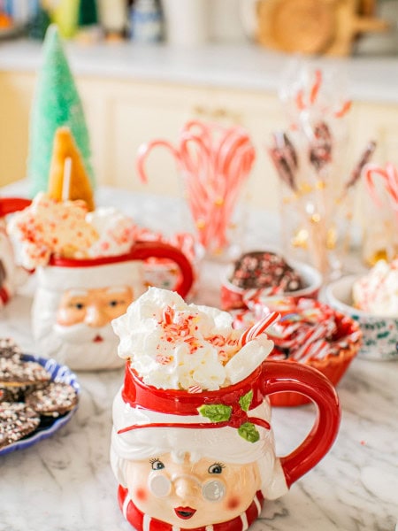 two mugs with santa and mrs. claus on them full of hot chocolate with toppings surrounding them on a hot chocolate bar