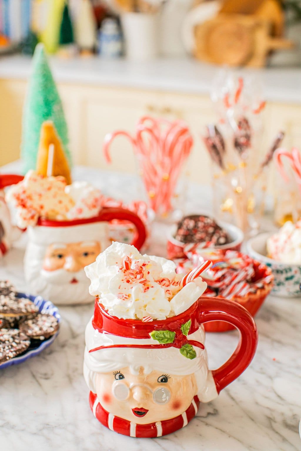 two mugs with santa and mrs. claus on them full of hot chocolate with toppings surrounding them on a hot chocolate bar