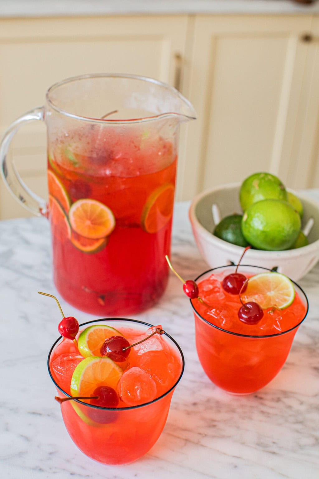 a large pitcher of dirty shirley cocktails beside two cocktail glasses full of the drink and a small bowl of extra limes