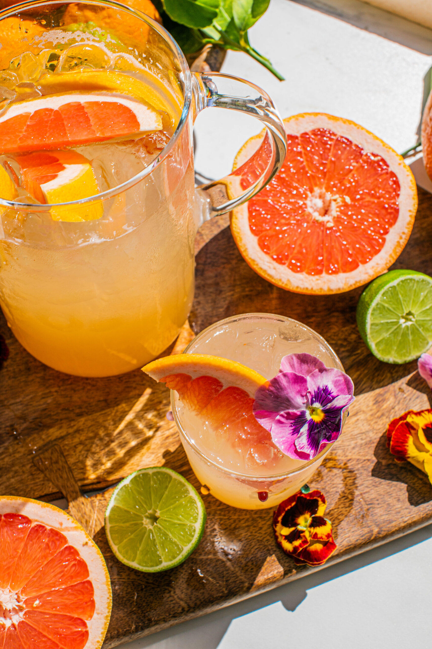 a pitcher of palomas beside a glass of the drink garnished with edible flowers and a grapefruit wedge