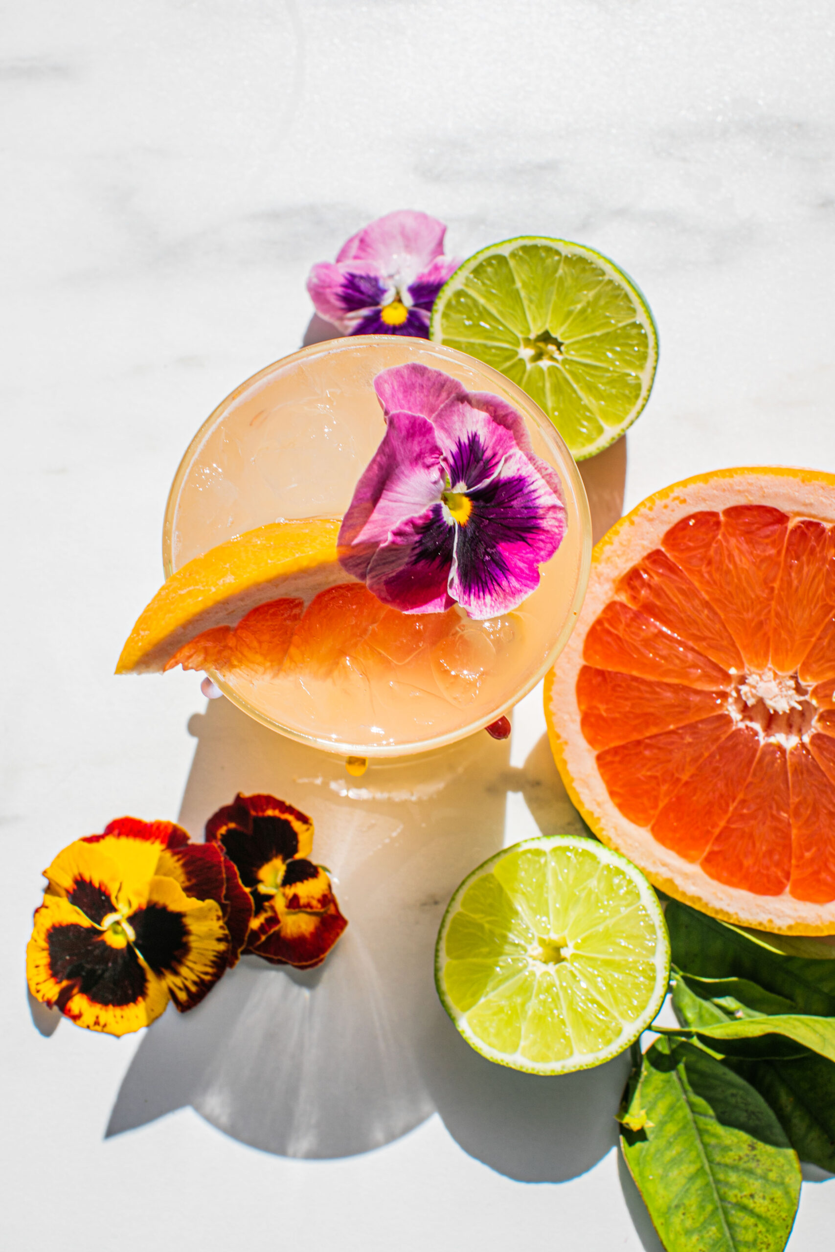 an overhead photo of a paloma garnished with edible flowers, a grapefruit wedge, and fresh lime and grapefruit scattered around it 