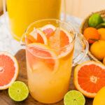 a large pitcher of grapefruit palomas with grapefruits and limes around it