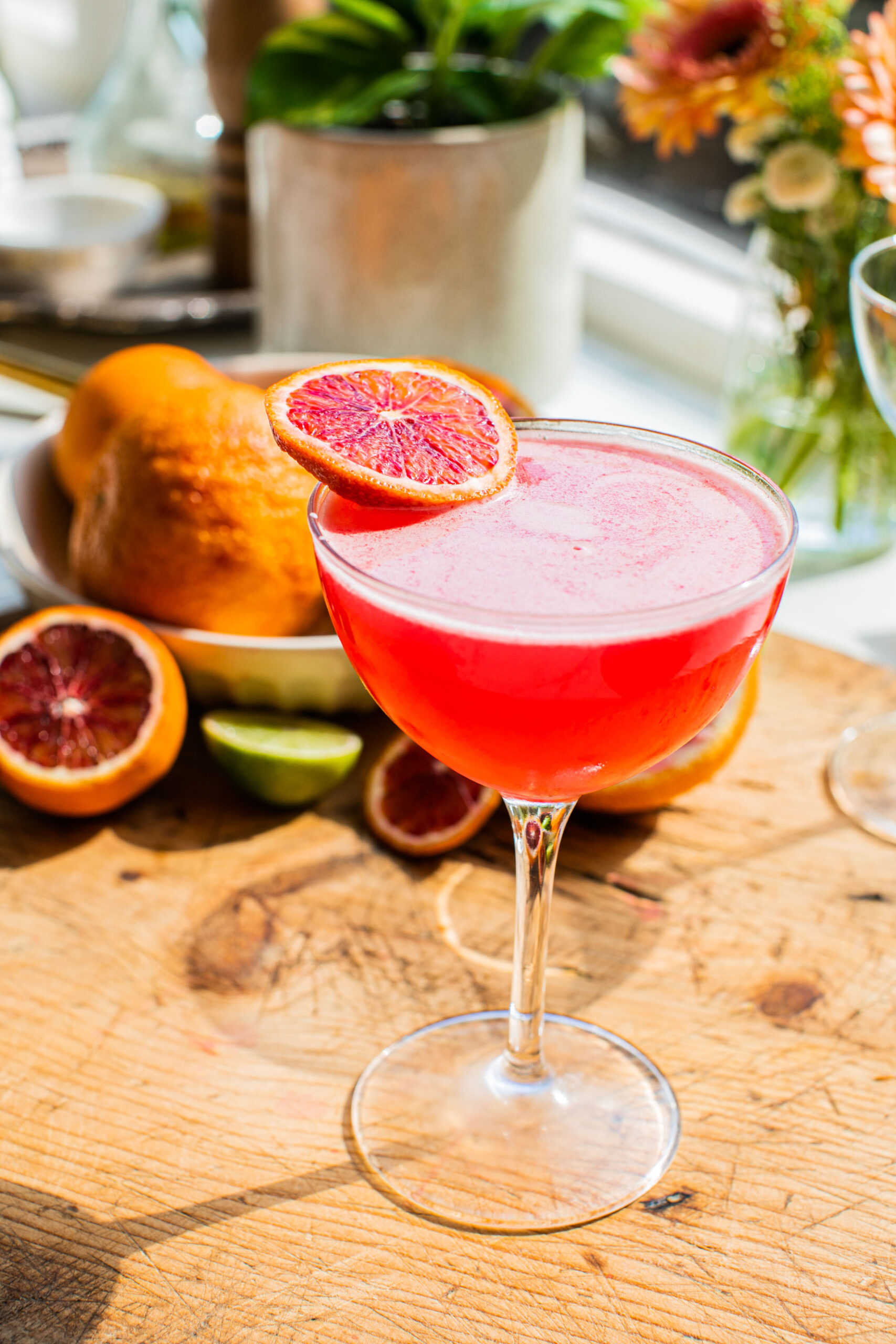 a blood orange martini garnished with an orange wheel and displayed in front of a bowl of oranges and limes