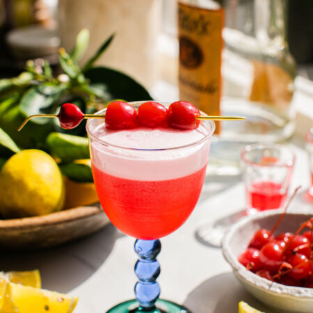 a glass of cherry vodka sour with vodka, cherries, and lemons in the background