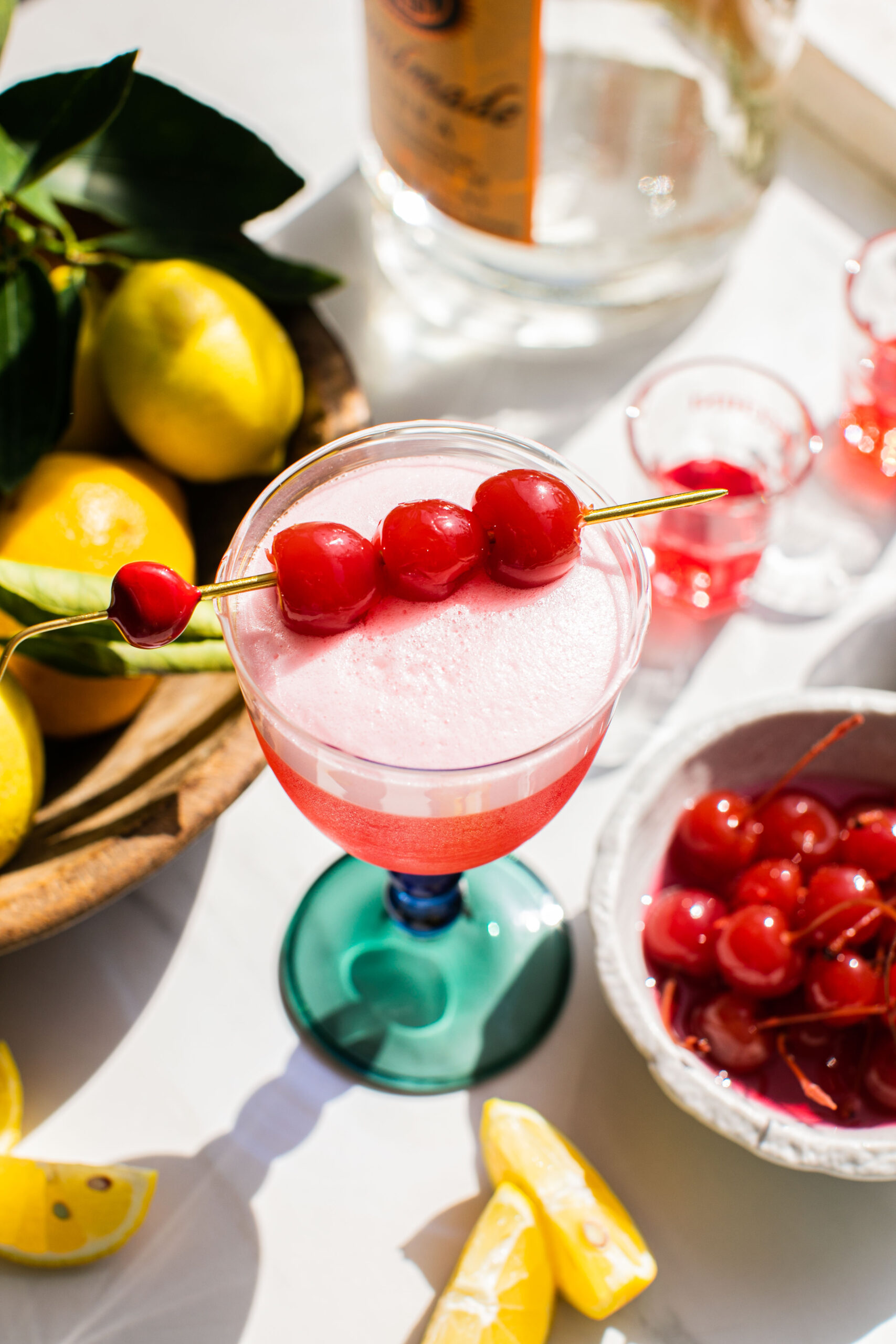 an overhead shot of a cherry vodka sour beside lemon wedges, cherries, and a vodka bottle