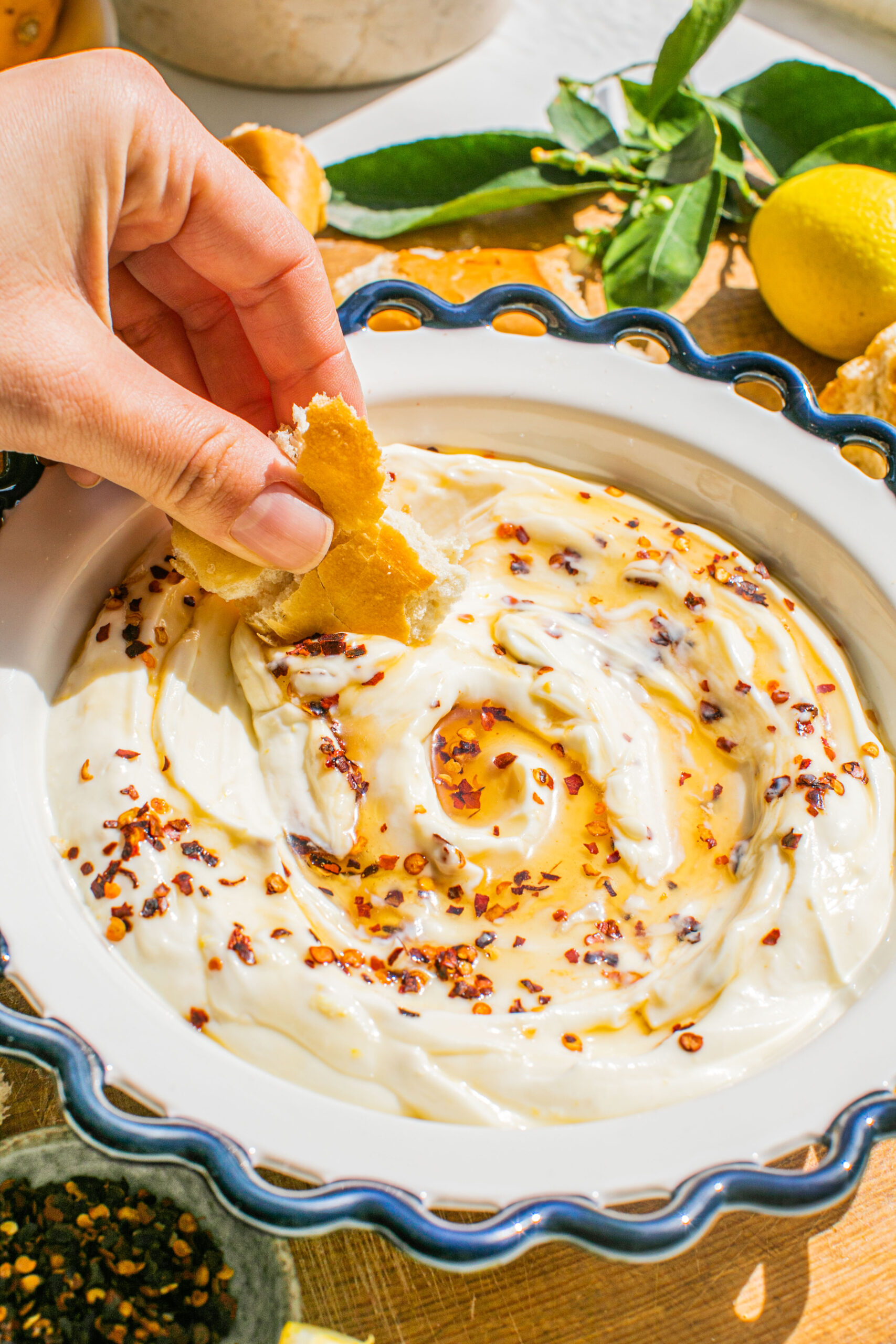 a close up photo of whipped ricotta in a bowl topped with hot honey and red pepper flakes and a hand dipping a piece of bread in it