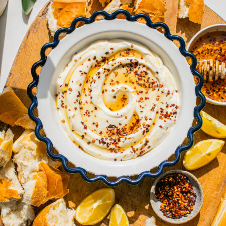 a bowl of whipped ricotta topped with hot honey and red pepper flakes, pictured with bread chunks and lemon slices on a wooden board around it