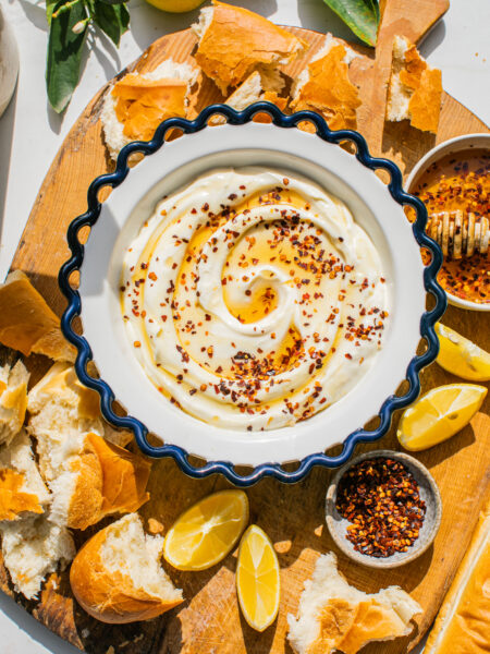 a bowl of whipped ricotta topped with hot honey and red pepper flakes, pictured with bread chunks and lemon slices on a wooden board around it