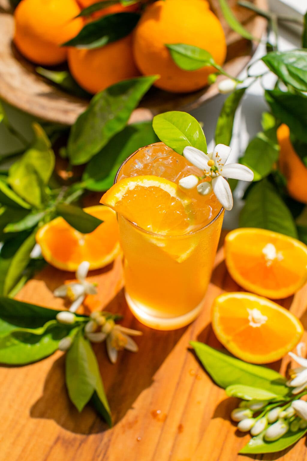 a glass of an orange gin and tonic with an orange blossom and orange slice as garnish, plus leaves and extra orange slices scattered around