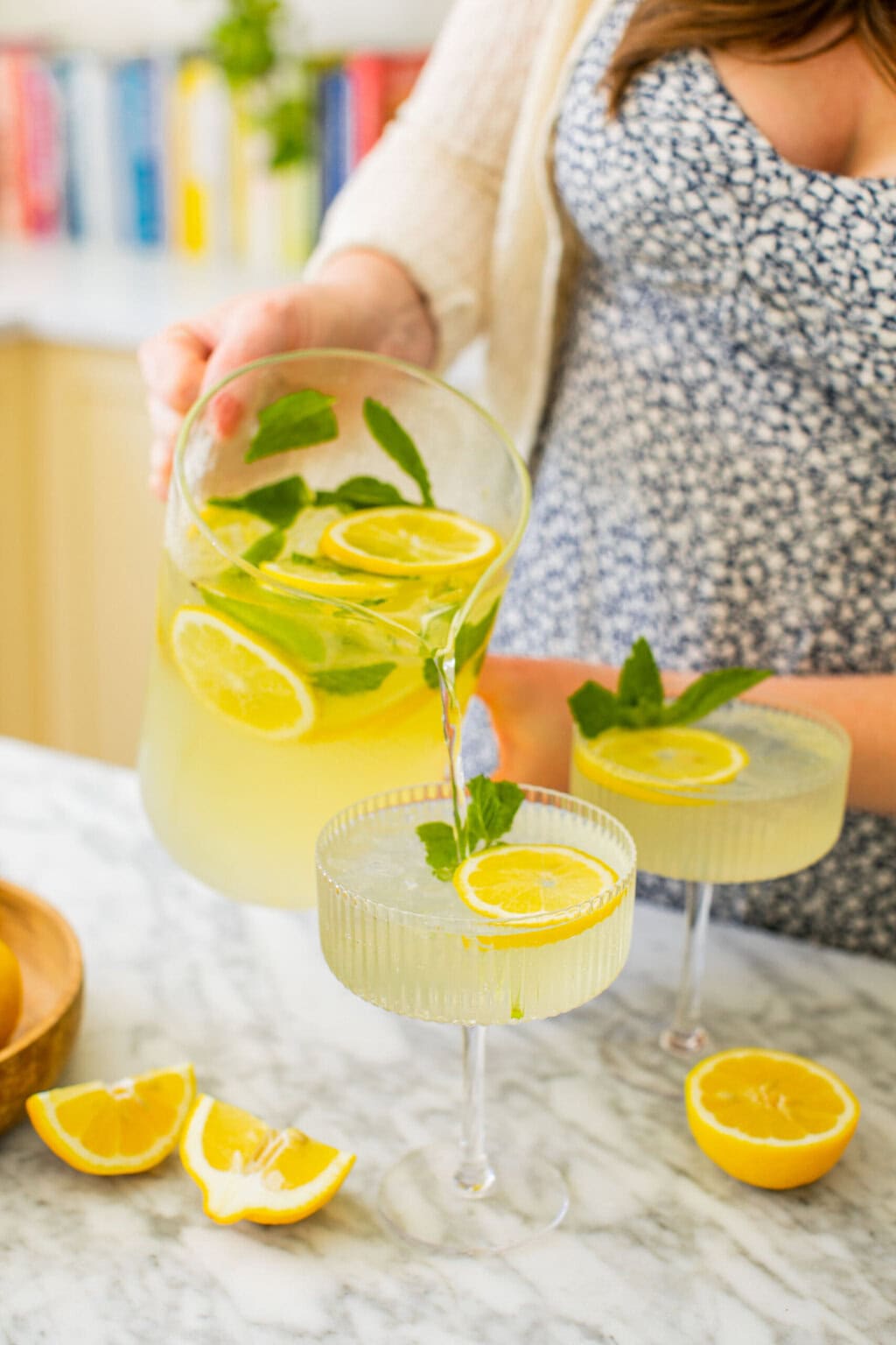 Elizabeth pouring the limoncello spritz from the pitcher into one of two coupe glasses garnished with lemon slices and mint leaves