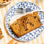 a slice of carrot loaf cake on a blue and white plate beside a fork with whole carrots on the table cloth around it