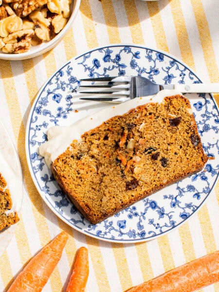 a slice of carrot loaf cake on a blue and white plate beside a fork with whole carrots on the table cloth around it