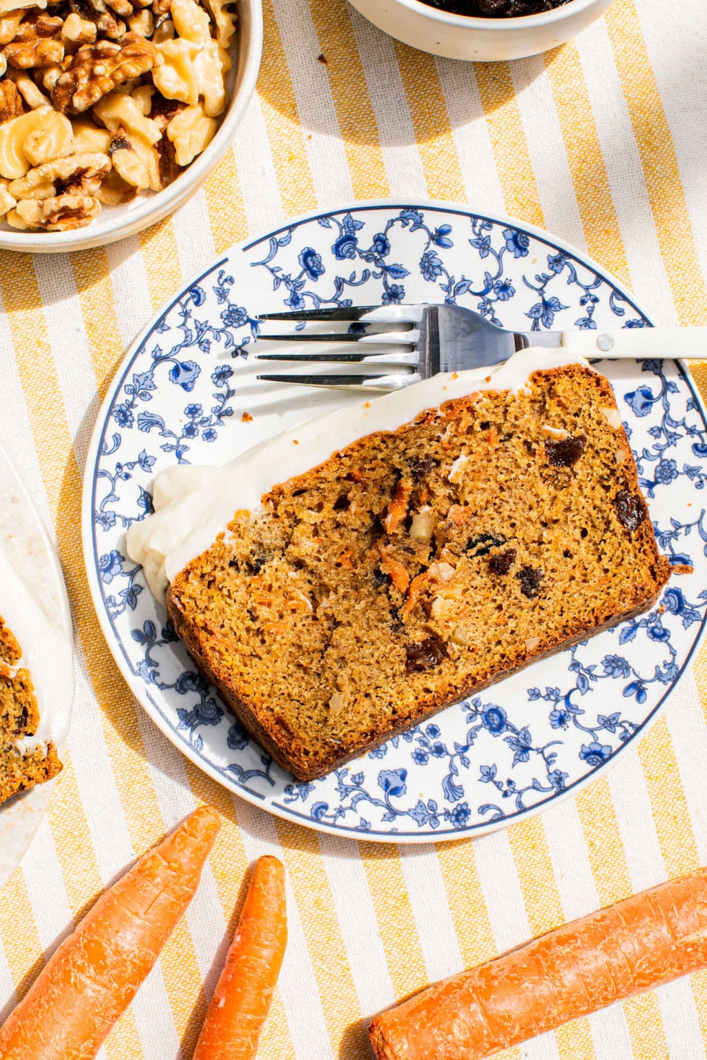 a slice of carrot loaf cake on a blue and white plate beside a fork with whole carrots on the table cloth around it