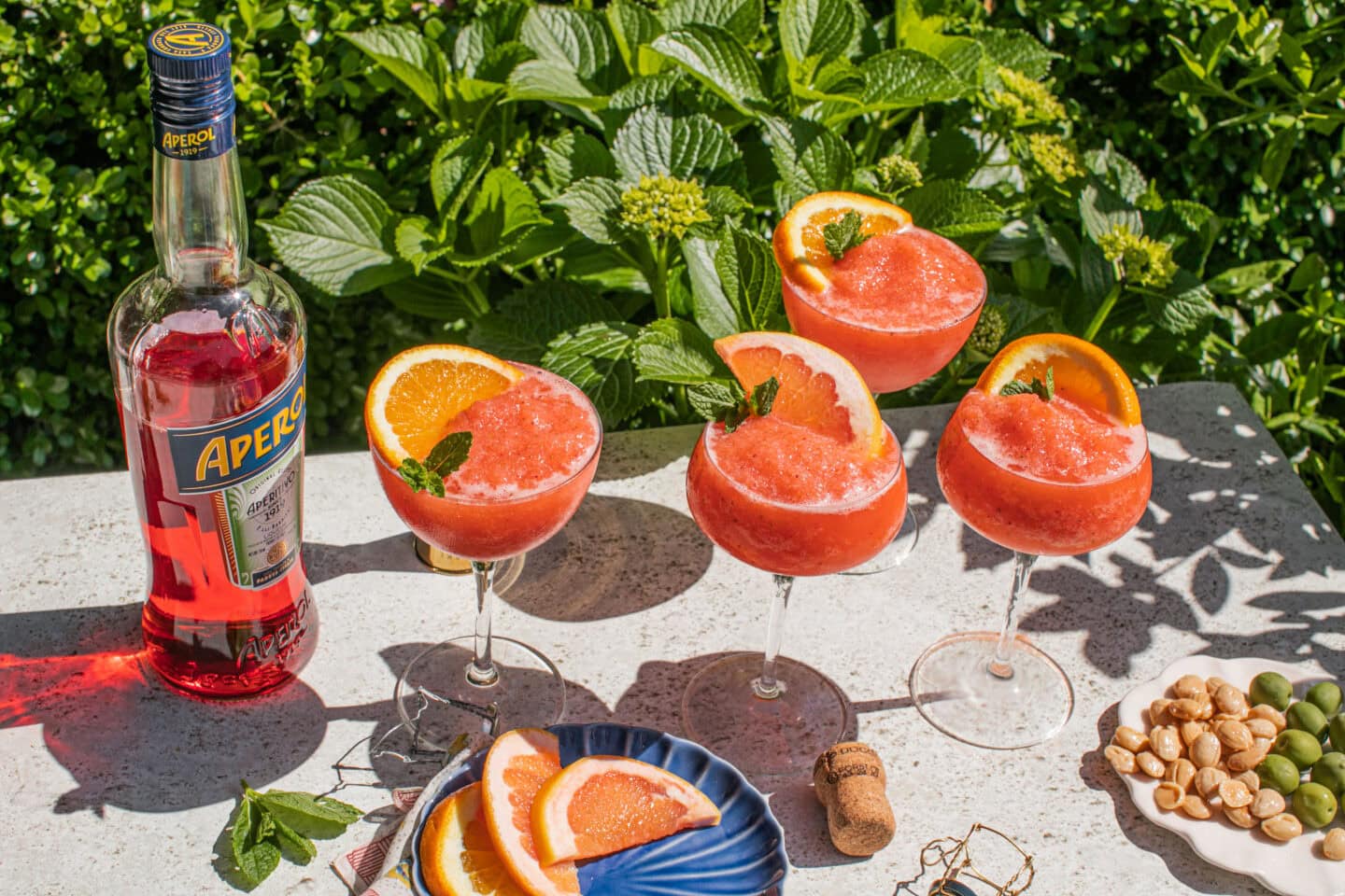 four frozen aperol spritz garnished with a grapefruit slice, fresh mint, a bottle of aperol, a plate of grapefruit and a plate of marcona almonds and green olives