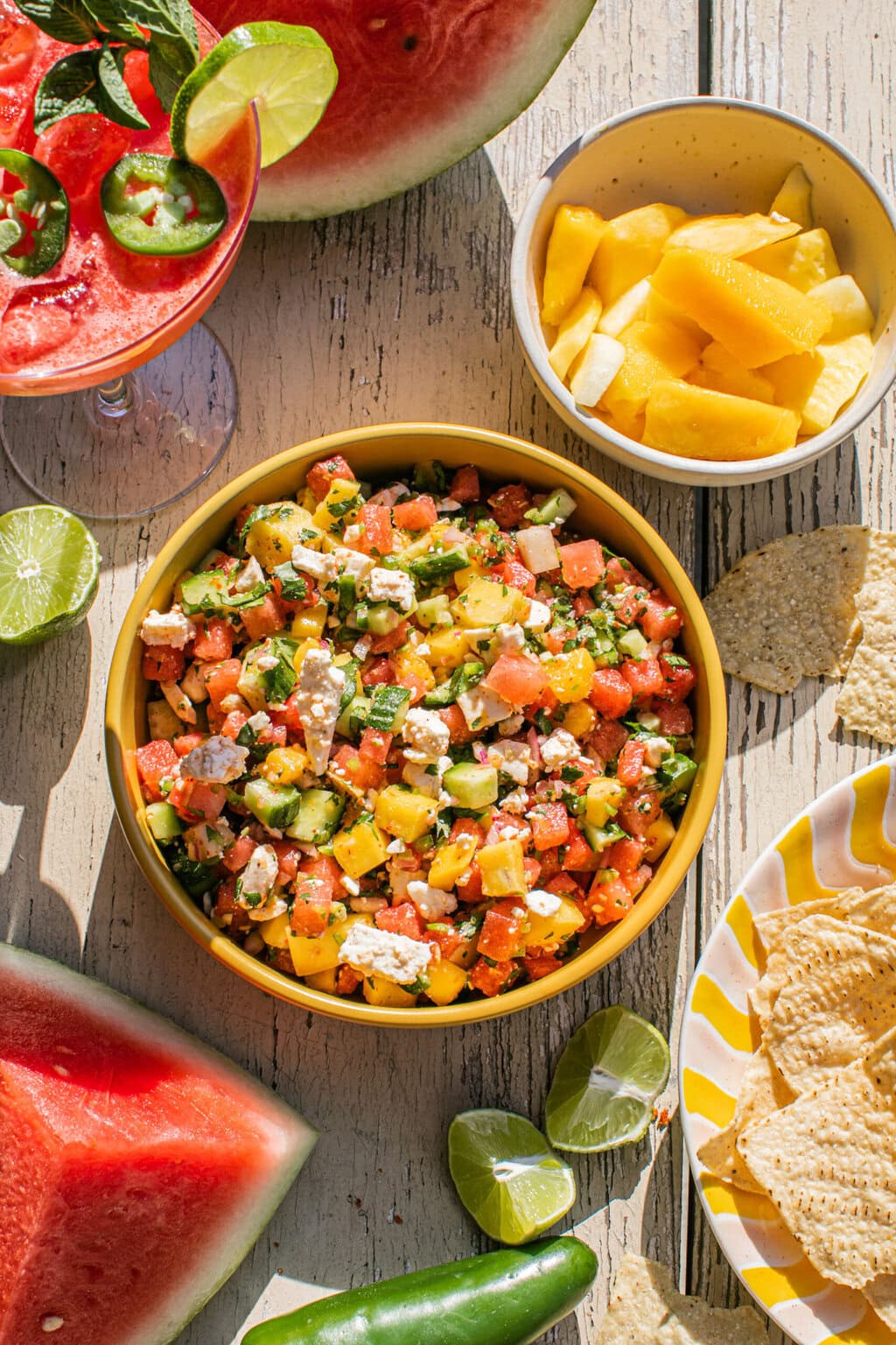 a bowl of watermelon feta salsa beside a bowl of more fruit, tortilla chips, and a watermelon cocktail