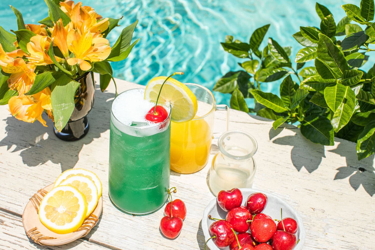 a blue lagoon cocktail beside lemons, cherries, pineapple juice, and lemonade, all poolside