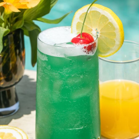 a blue lagoon cocktail beside pineapple juice and lemonade, garnished with a lemon slice and cherry