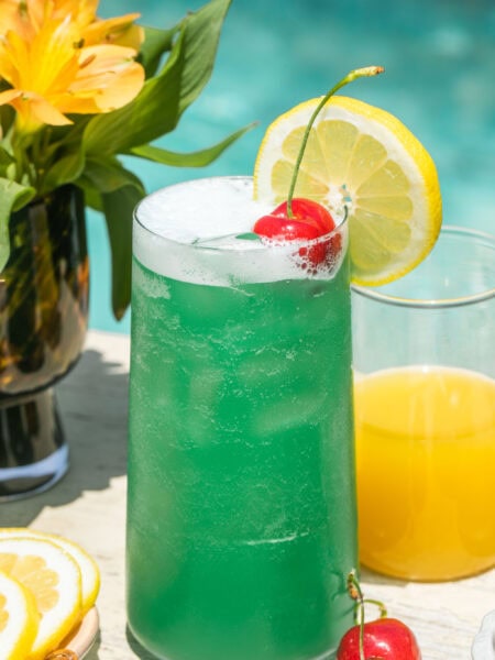 a blue lagoon cocktail beside pineapple juice and lemonade, garnished with a lemon slice and cherry