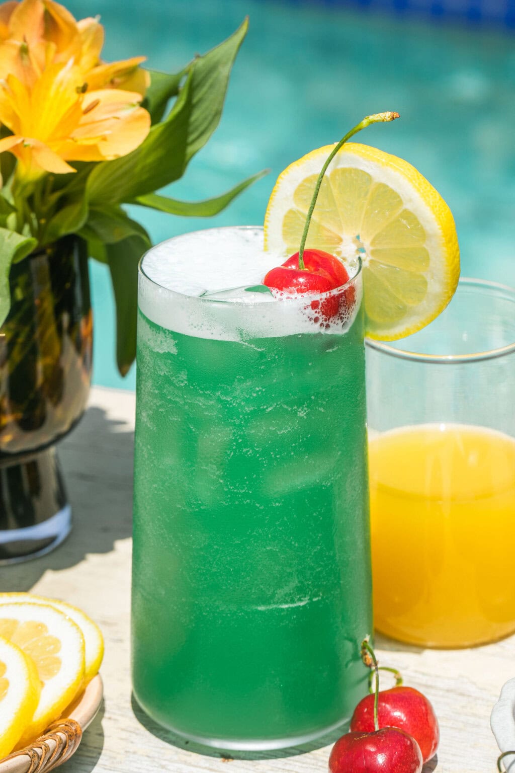 a blue lagoon cocktail beside pineapple juice and lemonade, garnished with a lemon slice and cherry