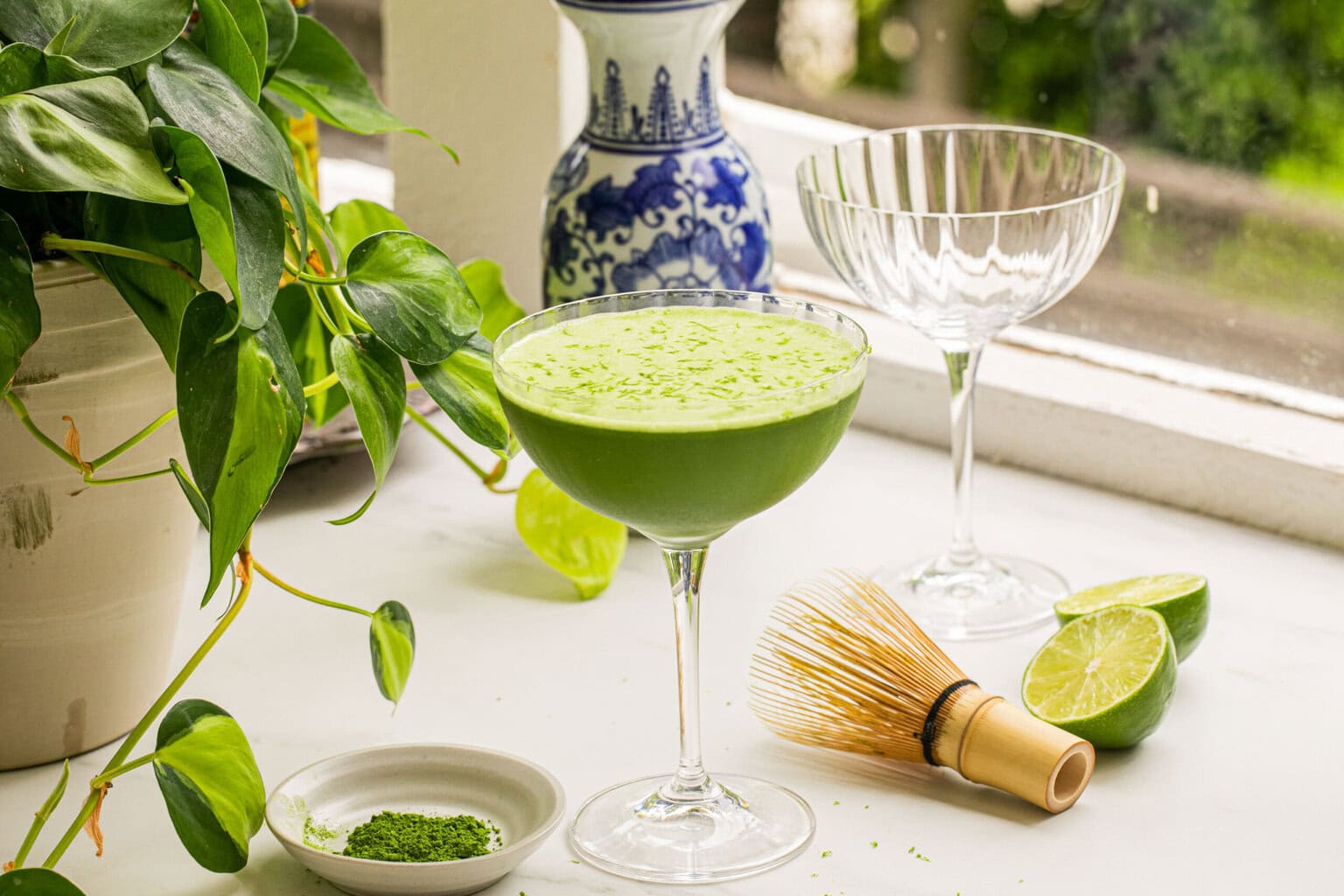 a matcha martini pictured beside matcha powder and a matcha whisk with limes to the right and a plant to the left