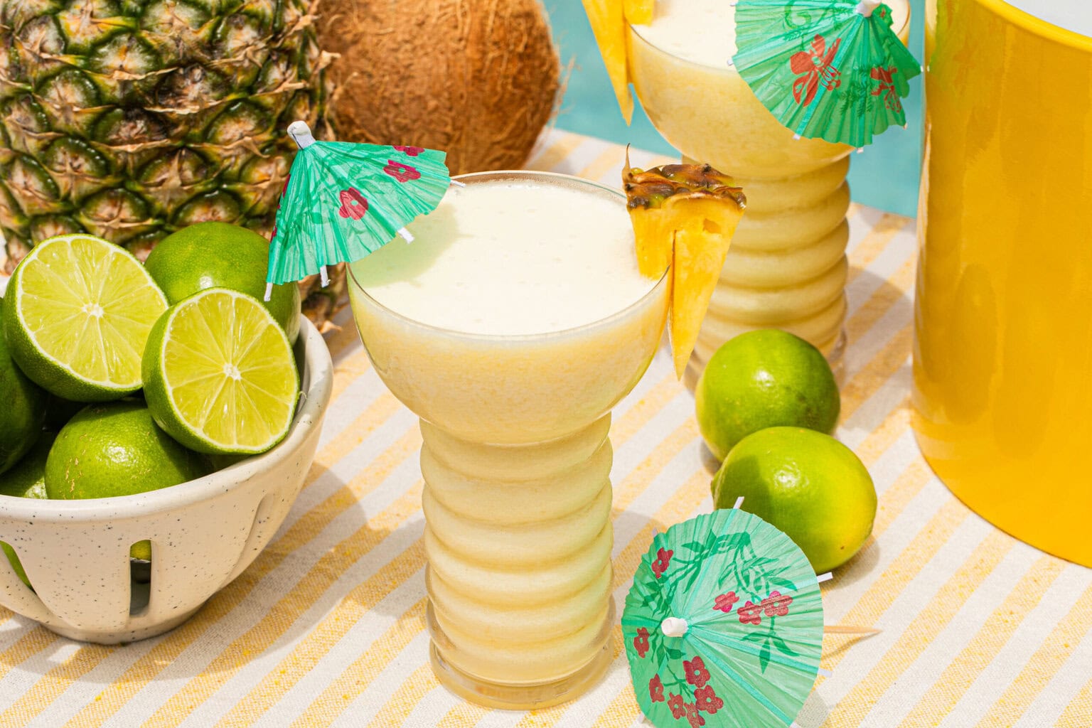 a mocktail with pineapple for garnish and a mini umbrella beside limes, a pineapple, and a coconut