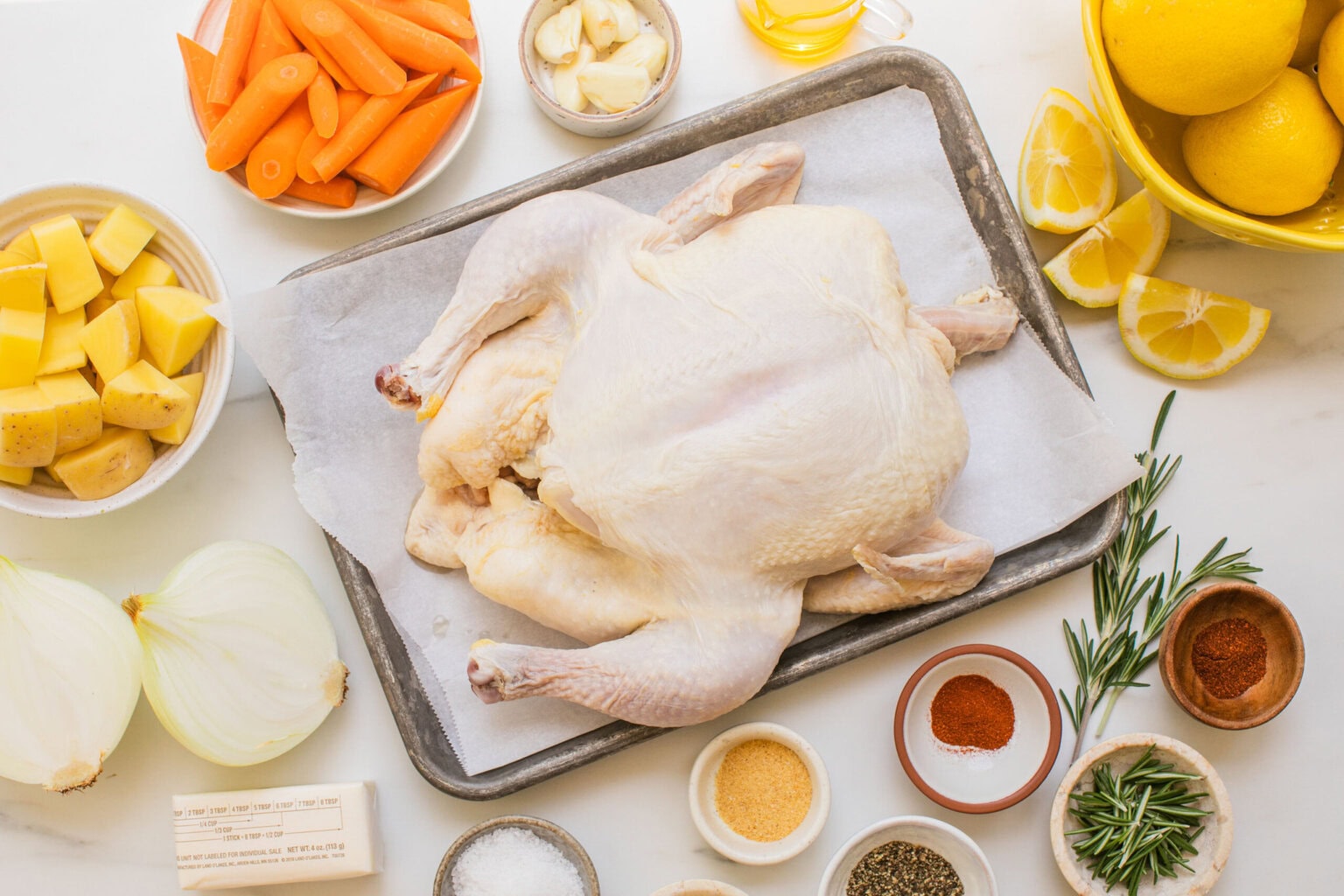 top shot of a whole raw chicken on. a sheet pan surrounded by ingredients like potatoes, onions, lemon slices, butter, and seasonings