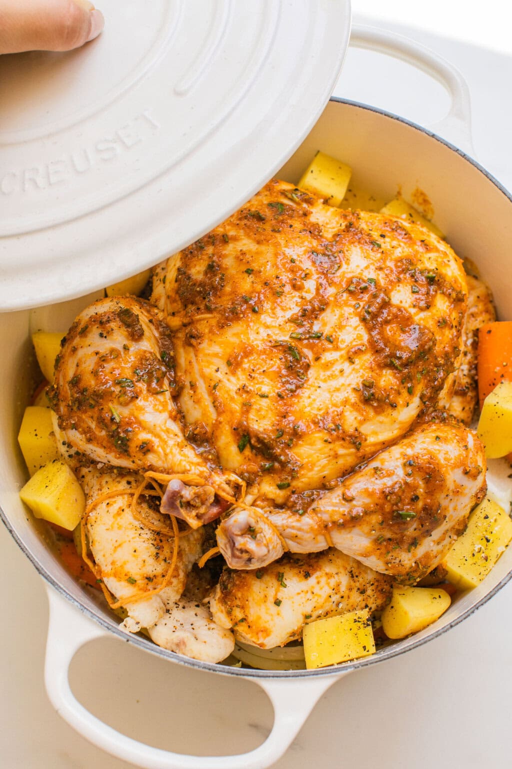 an overhead shot of a whole chicken in a dutch oven coated with seasoning on top of potatoes and veggies