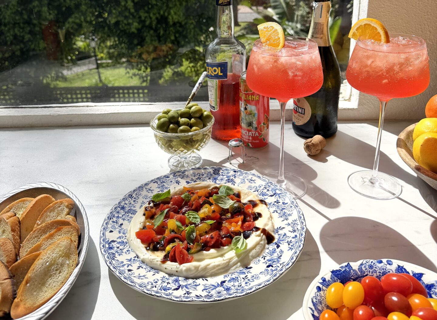 crostini slices in a silver platter, a blue and white platter with whipped ricotta and bruschetta topper, two large wine glasses filled with aperol spritz