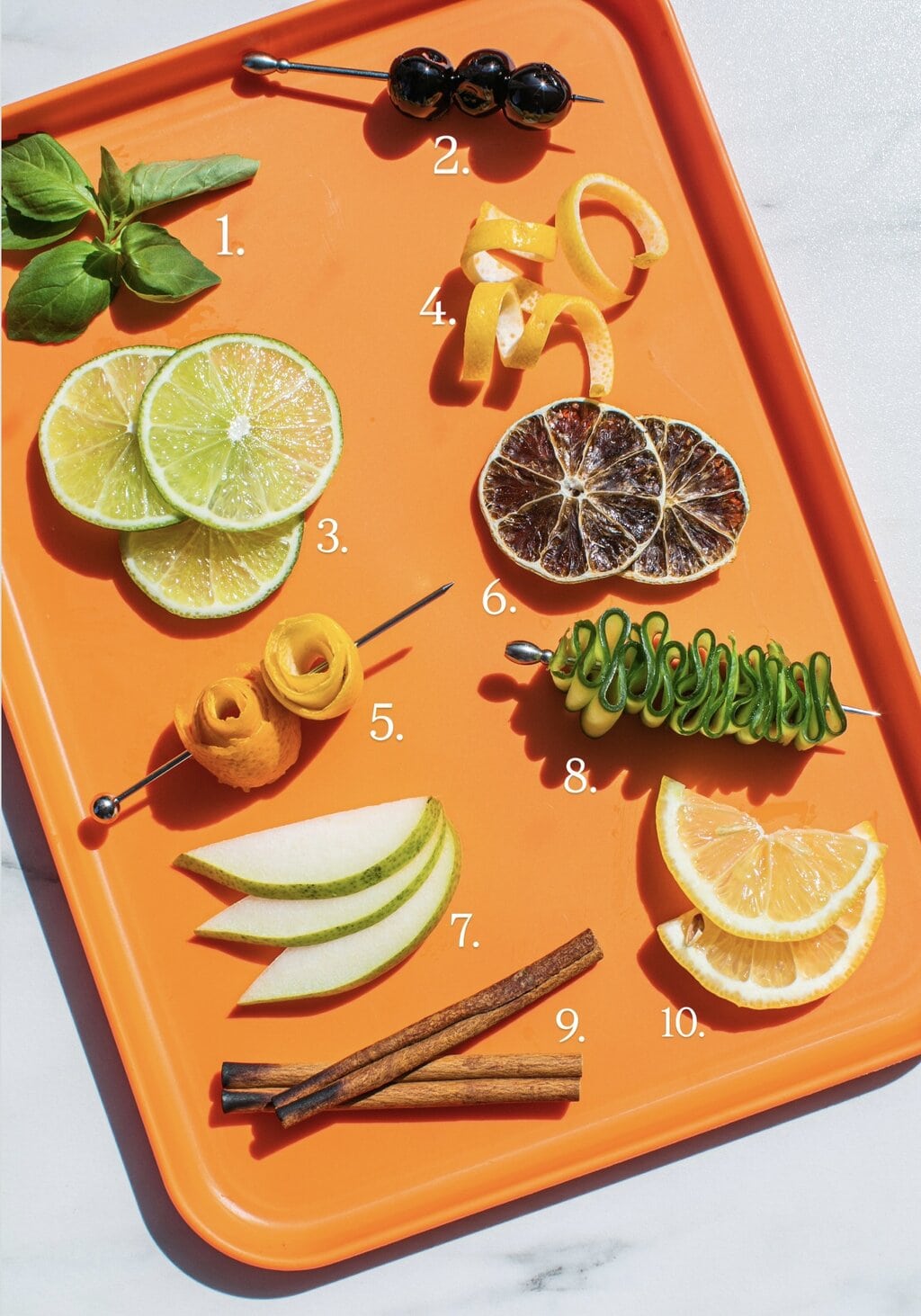 orange tray filled with cocktail garnishes like basil bouquet, luxardo cherries on a skewer, lime wheels, lemon twists, orange rosettes, dehydrated limes, pear slices, cucumber ribbons, cinnamon sticks and lemon half moons