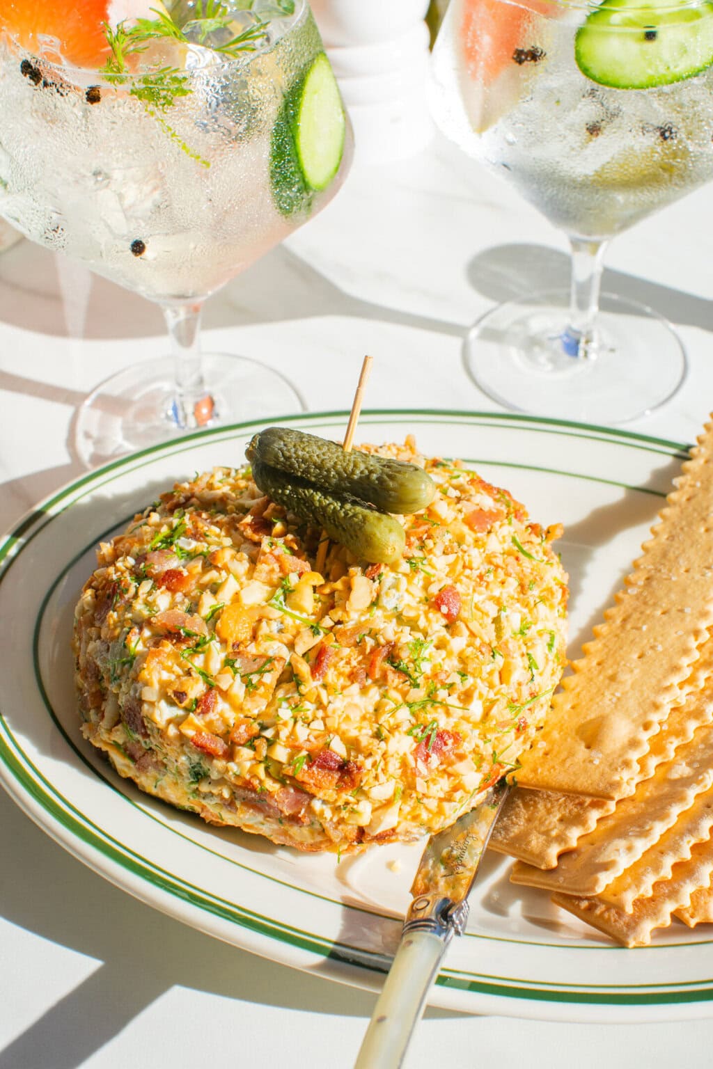 dill pickle cheese ball on a green and white platter served with crackers. two glasses filled with tonic water, grapefruit and cucumber