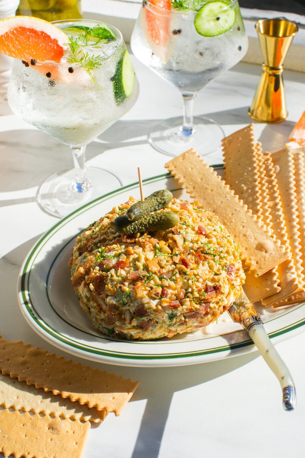 dill pickle cheese ball on a green and white platter served with crackers. two glasses filled with tonic water, grapefruit and cucumber