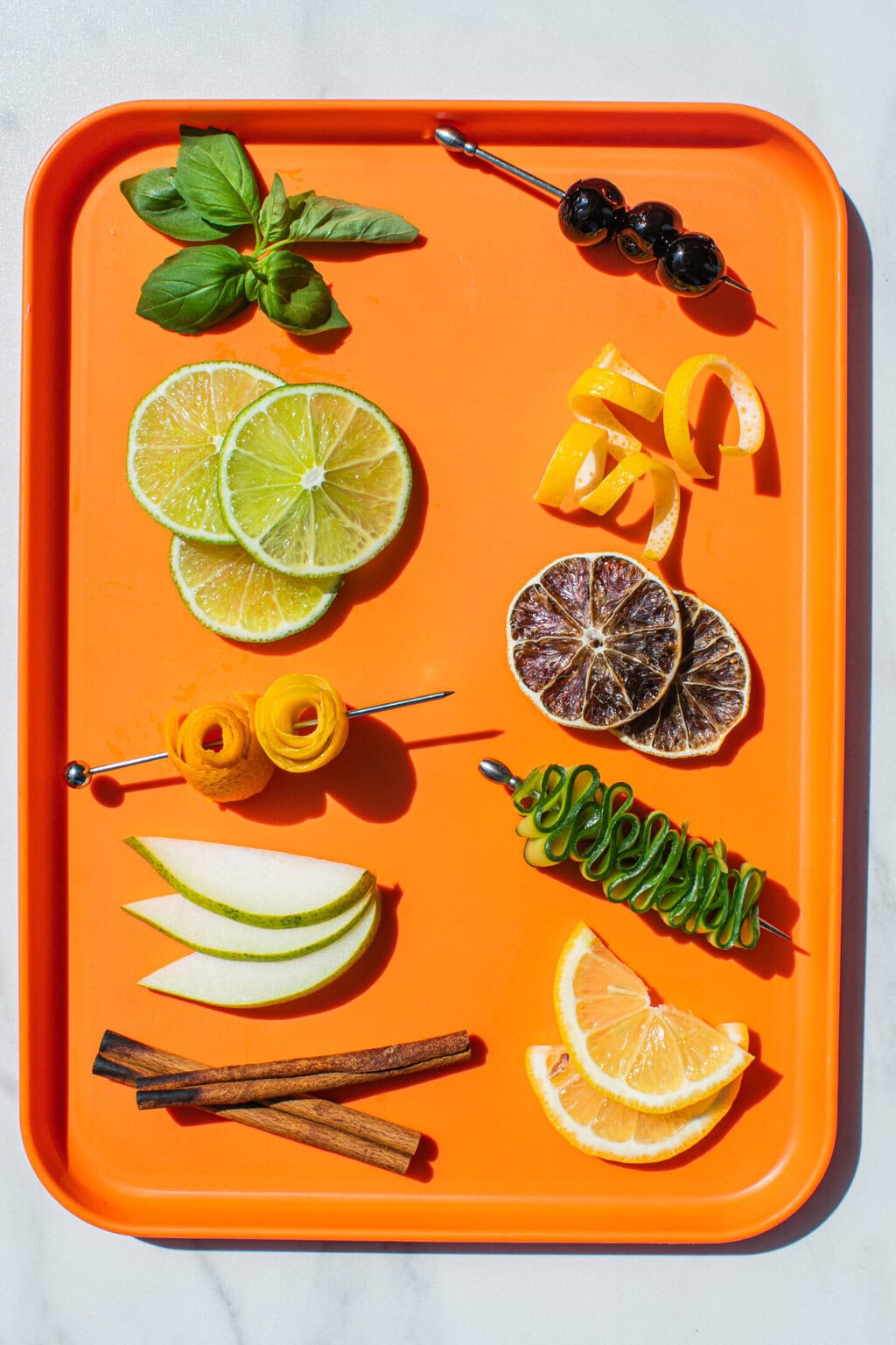 orange tray filled with cocktail garnishes like basil bouquet, luxardo cherries on a skewer, lime wheels, lemon twists, orange rosettes, dehydrated limes, pear slices, cucumber ribbons, cinnamon sticks and lemon half moons