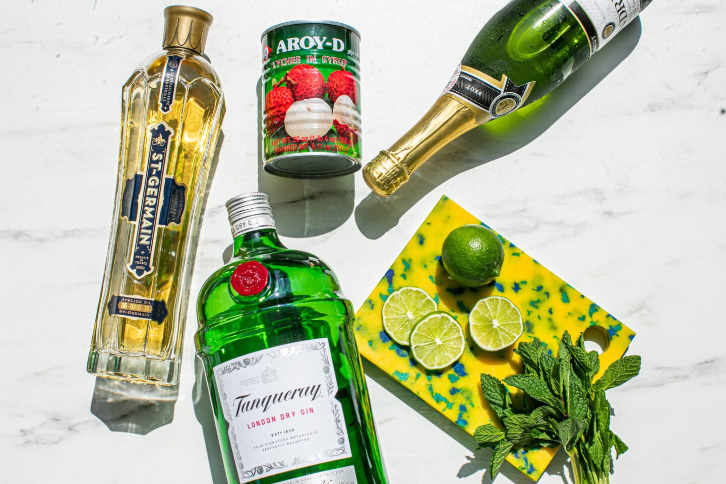 bottle of st germain elderflower liqueur, gin bottle, can of lychees, lime wheels, mint sprigs and a bottle of prosecco