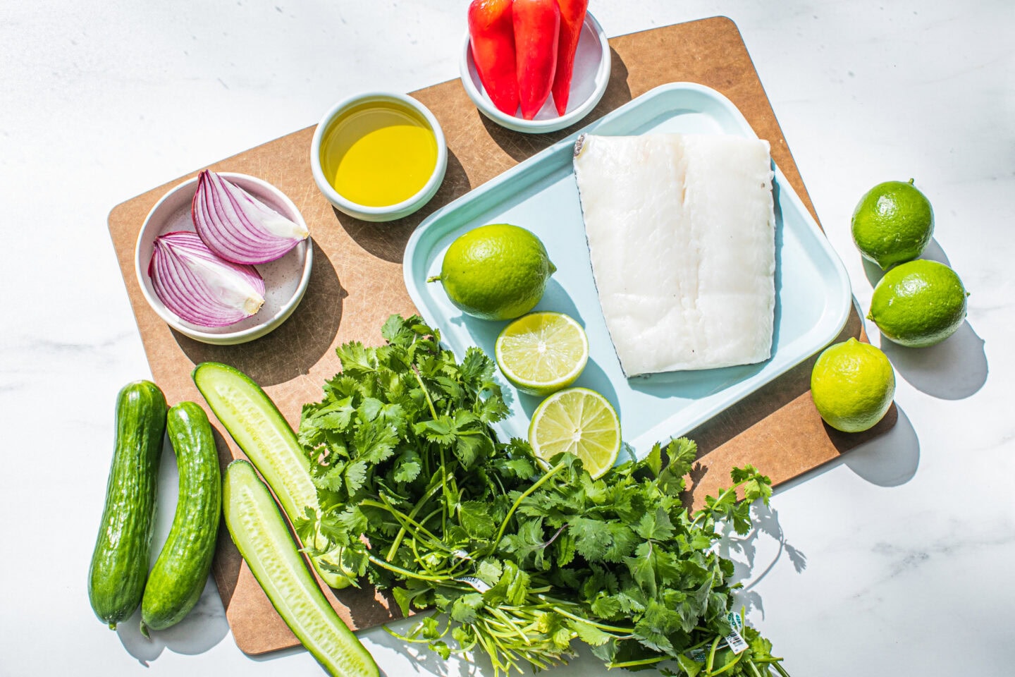 red onion, cucumbers, olive oil bowl, cilantro, limes, sea bass piece on a wooden cutting board