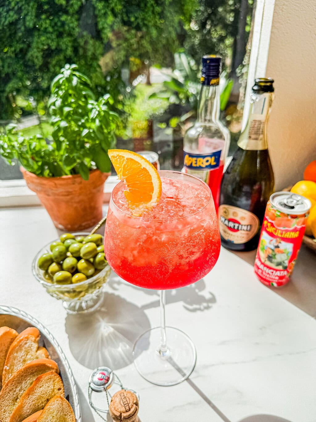 a small bowl of green olives, a wine glass filled with aperol spritz, a can of italian soda, a bottle of aperol, a bottle of prosecco