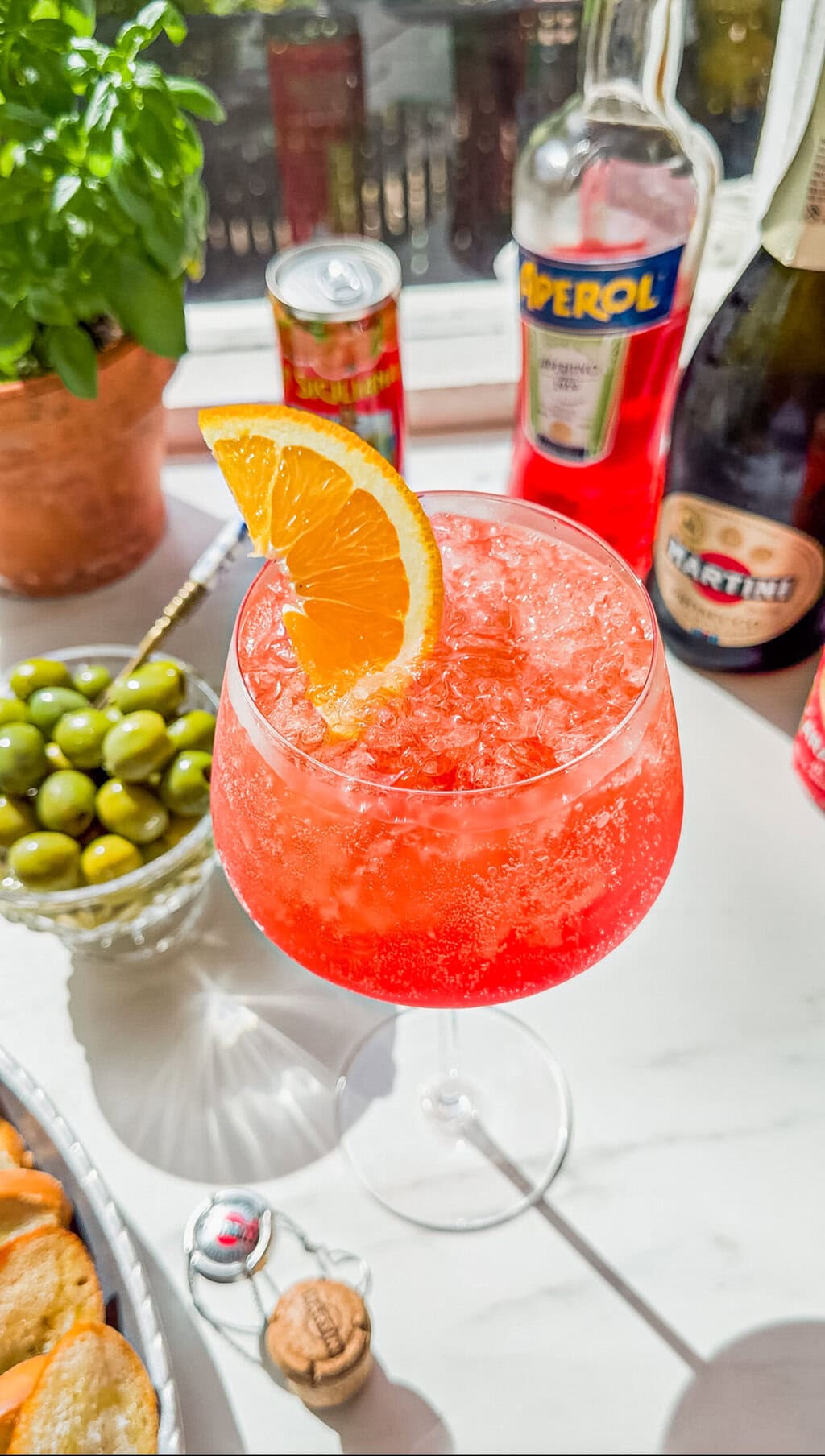 a small bowl of green olives, a wine glass filled with aperol spritz, a can of italian soda, a bottle of aperol, a bottle of prosecco