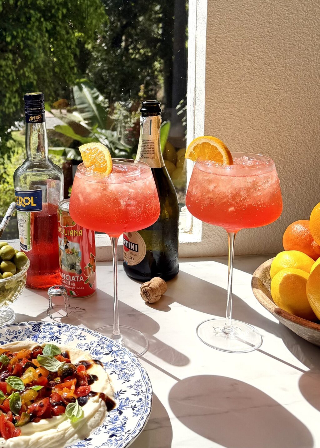 two glasses of aperol spritz on a counter with a bottle of aperol, prosecco and italian soda