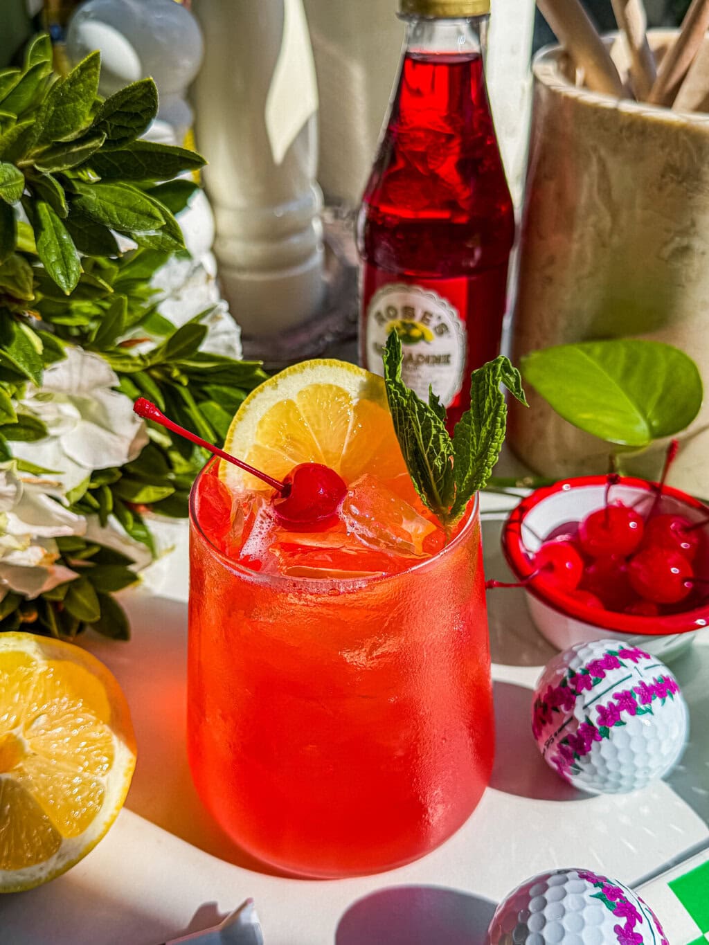 The azalea cocktail garnished with a lemon wheel, maraschino cherry and mint sprig. Azalea flowers and a bottle of grenadine. golf balls and a small bowl of maraschino cherries