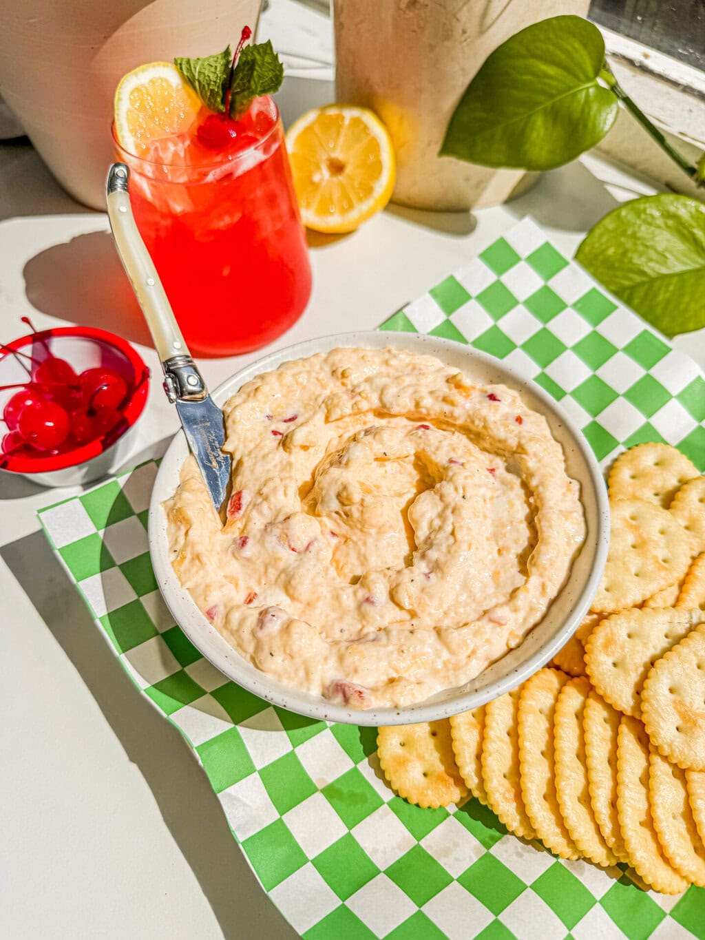a white bowl of the dip with a white spreader and crackers on a green and white checkered paper liner