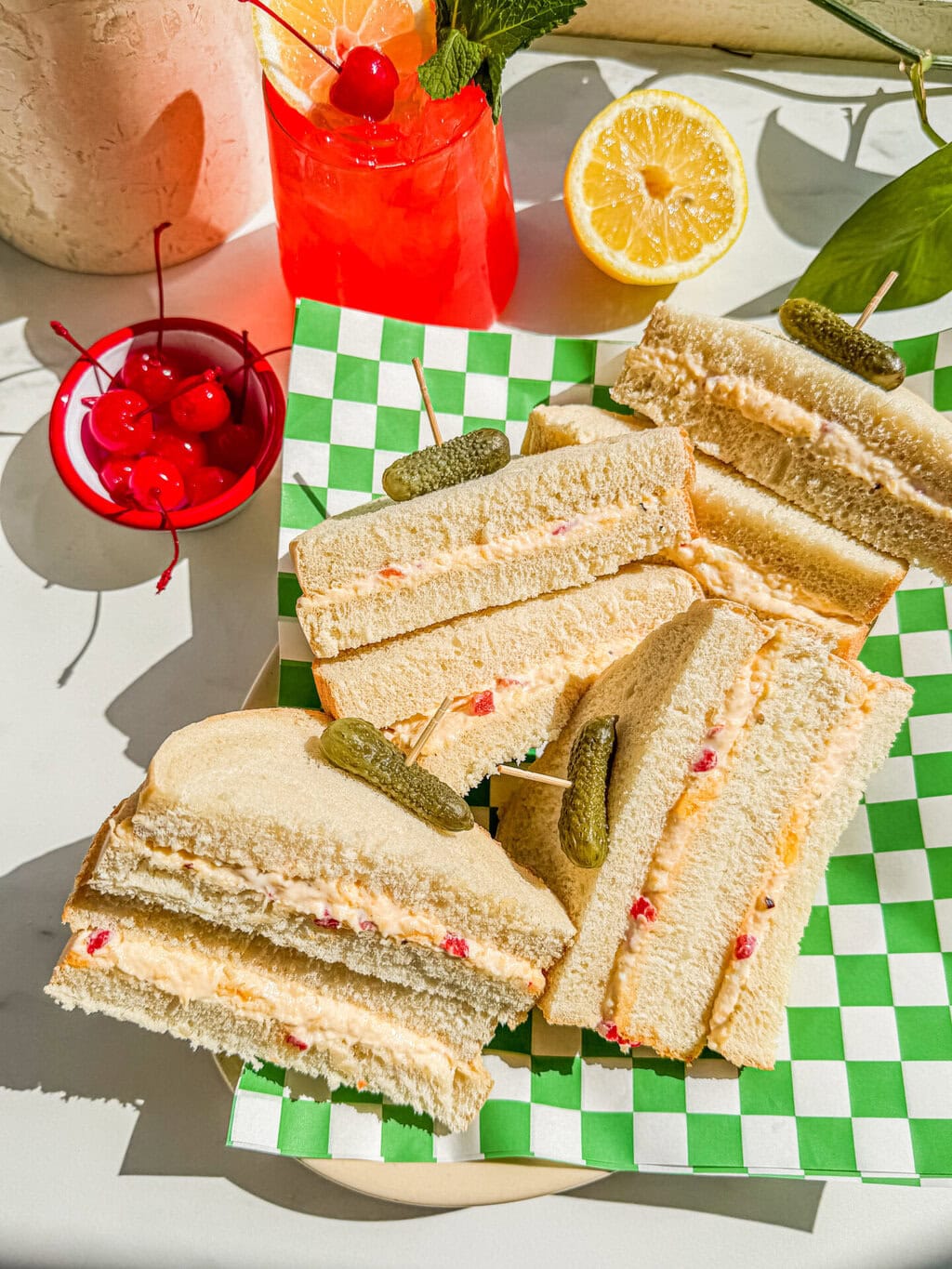 a few slices of the sandwiches with a cornichon garnish and an azalea cocktail on a green and white checkered wrapper