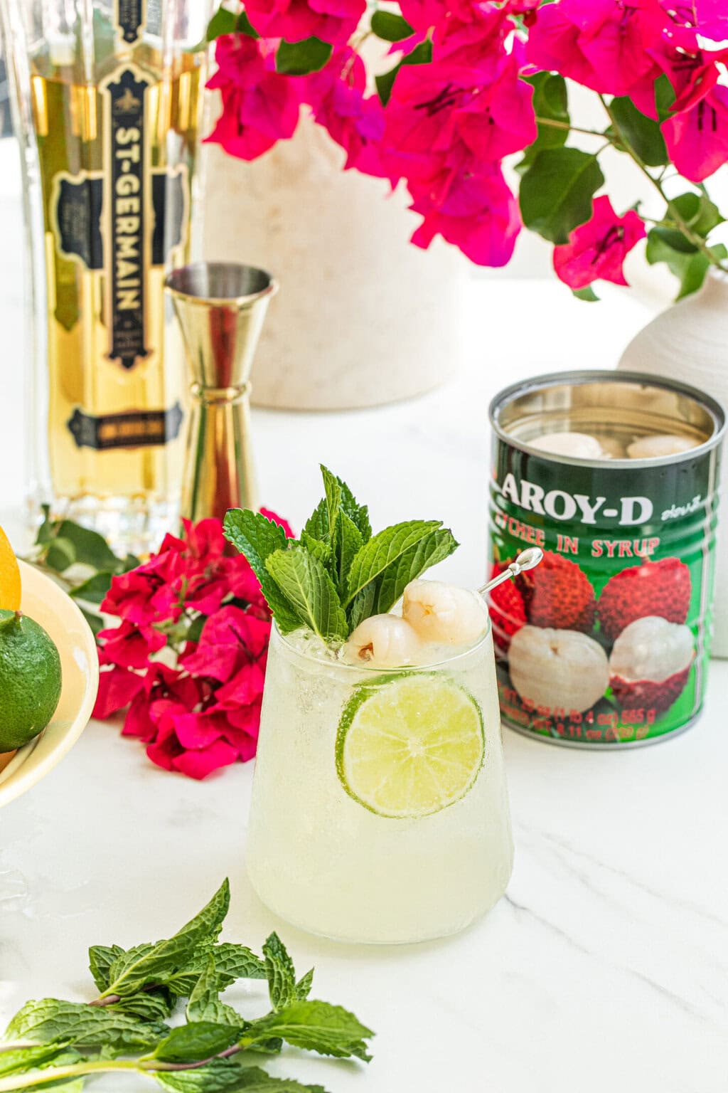 a stemless wine glass filled with lychee cocktail spritz, garnished with two lychees, a lime wheel, fresh mint, can of lychees, purple plant in the background, bottle of elder flower liqueur and a gold jigger