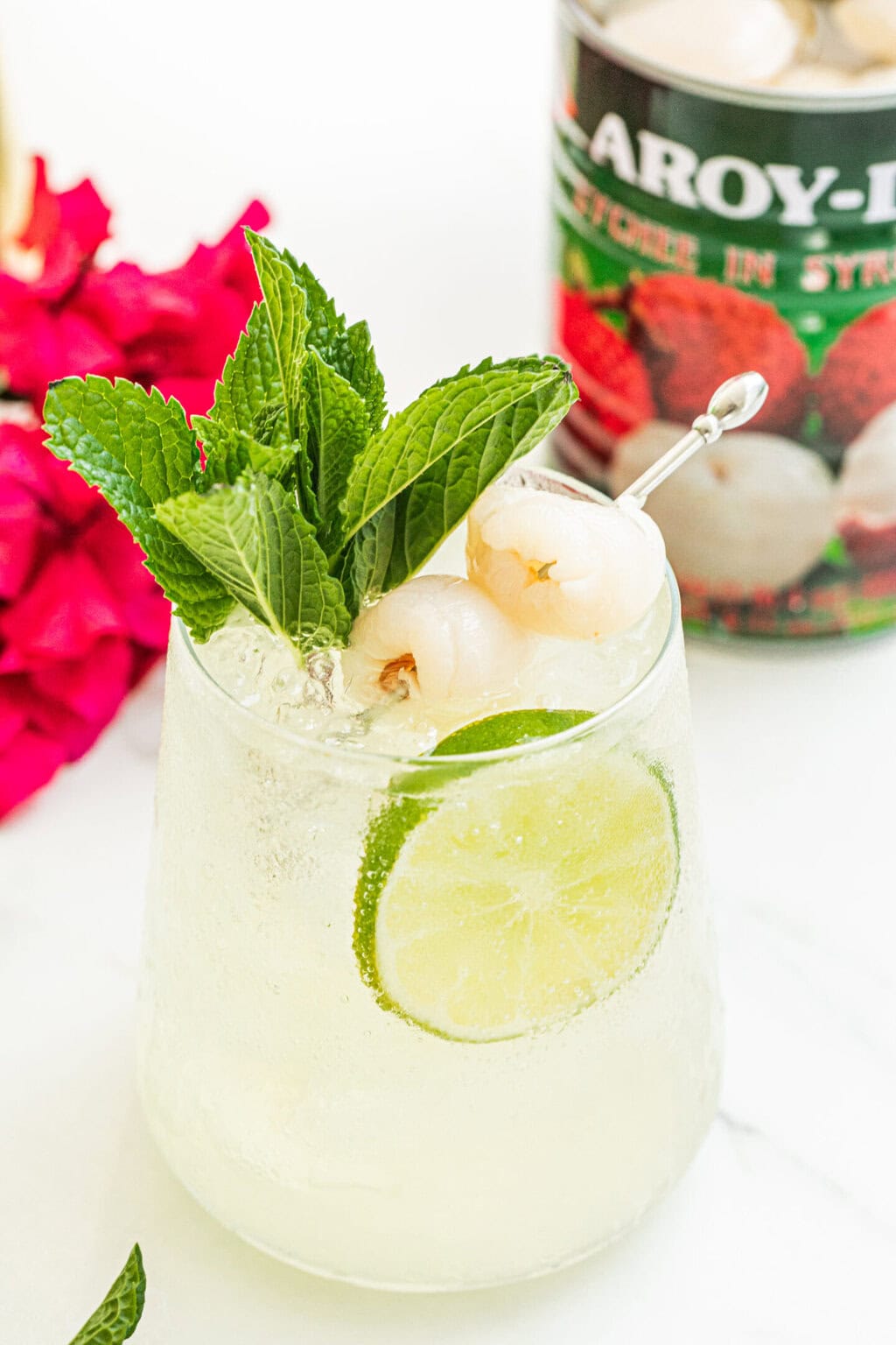 a stemless wine glass filled with lychee cocktail spritz, garnished with two lychees, a lime wheel, fresh mint, can of lychees, purple plant in the background 