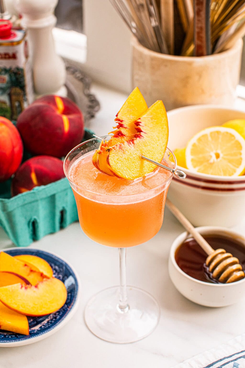 a basket of peaches, a bowl of lemons, the peach martini, peach slices and a small bowl of honey