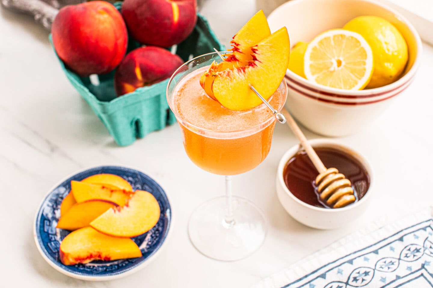 peach slices in a small blue dish, whole peaches in a blue basket, a peach martini recipe garnished with peach slices, a bowl of honey and a bowl of lemons