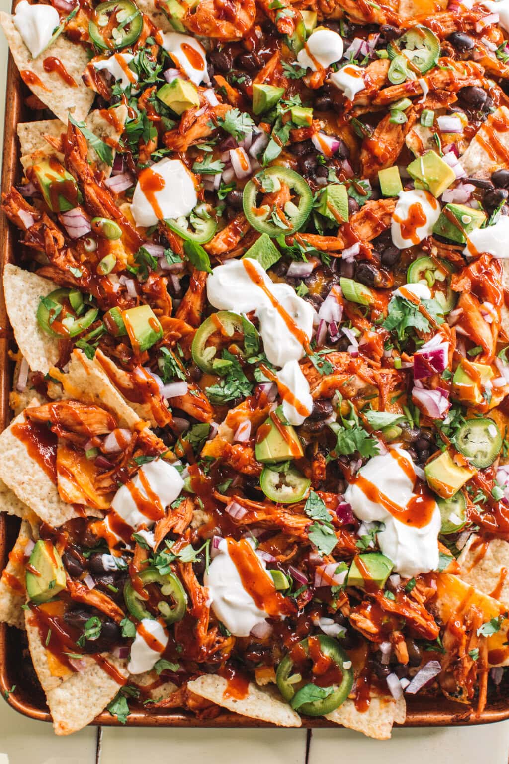 a platter of saucy bbq chicken nachos topped with bbq sauce, red onion, avocado, jalapeno slices and sour cream dollops 